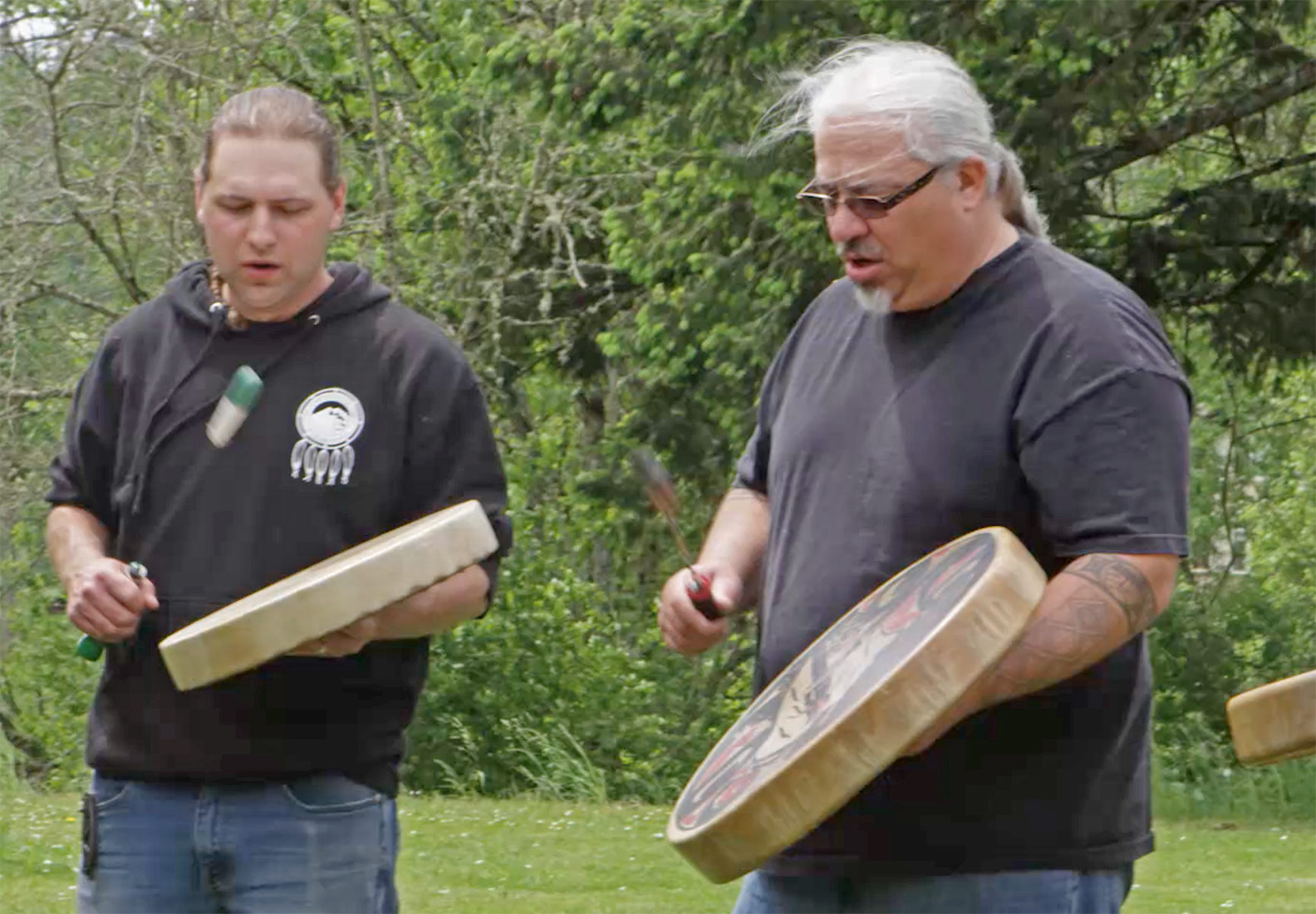 Two members of the Confederated Tribes of Grand Ronde beating drums in an outdoor ceremony.